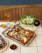 A high-quality, well-composed flat-lay photograph of a deep-dish pizza meal, accompanied by a fresh salad, dipping sauces, and glasses of wine, set against a clean, warm-toned wooden background.