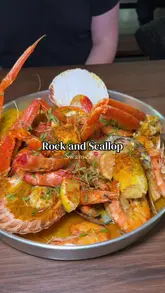 A creator visits Rock and Scallop at Albert Hall in Swansea to try their seafood boil. The video features close-ups of the large seafood platter, including lobster, crab, prawns, and scallops, along with the creator's reaction to the food and the experience of eating it with a bib and gloves.