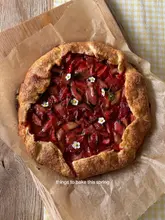 A high-quality, overhead shot of a freshly baked fruit galette, likely strawberry and rhubarb, garnished with small white flowers. The pastry is golden brown and the fruit filling looks glossy and vibrant, presented on parchment paper atop a rustic wooden board.