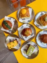A vibrant, high-quality overhead flat-lay photograph showcasing a variety of baked goods from Cantoast Bakery, arranged on individual metal plates with branded parchment paper on a bright yellow table.