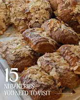 A high-quality, close-up photograph of a pile of almond croissants, serving as the background for a listicle-style graphic promoting bakeries in Northern Ireland. The image focuses on the texture and golden-brown colour of the pastries, making them look fresh and inviting.