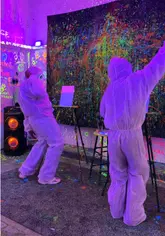A high-energy, fast-paced promotional video showcasing an activity venue called Splash House in Milton Keynes. The video features two creators in protective suits engaging in a neon paint fight and painting on canvases under UV lights, effectively demonstrating the fun and messy nature of the experience.