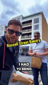 Two creators review a bakery near Croke Park, sampling a spicy chicken focaccia sandwich and a ham and cheese croissant. They discuss the freshness of the bread and the quality of the ingredients while standing outside the venue.