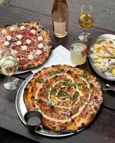 A flat-lay photograph of a table spread featuring two pizzas, a side dish of crisps, a bottle of rose wine, and a cocktail, set on a dark wooden outdoor table.