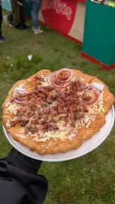 A first-person perspective shot of a freshly prepared Hungarian lángos, topped with sour cream, grated cheese, bacon, and red onion, held against a blurred outdoor festival background.