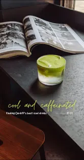 A high-quality, atmospheric shot of a coconut water matcha drink served in a glass, placed on a dark table alongside an open magazine. The lighting is moody and intentional, highlighting the vibrant green of the matcha against the dark background, creating a refreshing and aesthetic visual suitable for a cafe promotion.