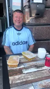 A man reviews Chinese takeaway food, specifically a crispy duck noodle box, chicken satay, and spring rolls, while sitting at an outdoor table.