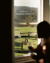 A high-contrast, atmospheric shot taken from inside a room looking out through a window at a sprawling, manicured countryside estate. The foreground features a silhouette of a person holding a glass of wine, creating a moody and aspirational 'getaway' aesthetic that effectively highlights the venue's scenic location.