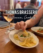A close-up shot of a hand dipping a crisp into a bowl of taramasalata at a restaurant table, with a wine glass and other diners visible in the background.