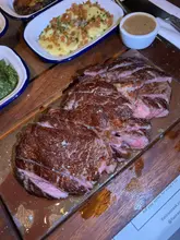 A top-down, slightly angled shot of a sliced ribeye steak served on a wooden board, accompanied by sides including mashed potatoes and creamed spinach, typical of a casual steakhouse dining experience.