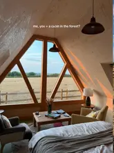 A beautifully composed, atmospheric shot of a cabin interior in Somerset, capturing the golden hour light and the scenic view through the large A-frame window. The image effectively communicates a sense of relaxation and escapism, perfectly aligning with the 'staycation' theme.