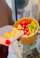 A vibrant, fast-paced video showcasing a new self-serve scoop bar called Mill & Grain. The creator demonstrates the process of building custom bowls with various toppings, including acai, cheesecake, and ice cream, highlighting the unlimited topping concept.