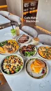 A high-quality, overhead flat-lay photograph showcasing a variety of dishes and drinks from Cafe Raha. The spread includes a breakfast bun, chicken caesar salad, lamb chops, chicken tikka loaded fries, a steak and onion toastie, and various specialty lattes and a cheesecake. The lighting is bright and even, highlighting the textures of the food against a clean marble tabletop.