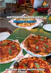 Two creators visit Gracey's Pizza in St Albans as part of their Hertfordshire food series. They show the exterior, the kitchen where dough is prepared, the menu, and enjoy a meal of olives, various pizzas, and cheesy garlic bread at an outdoor table.